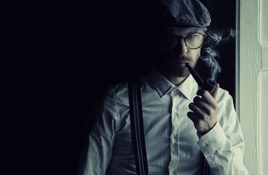 Man with beard wearing hat, glasses, white shirt with suspenders and smoking a pipe.