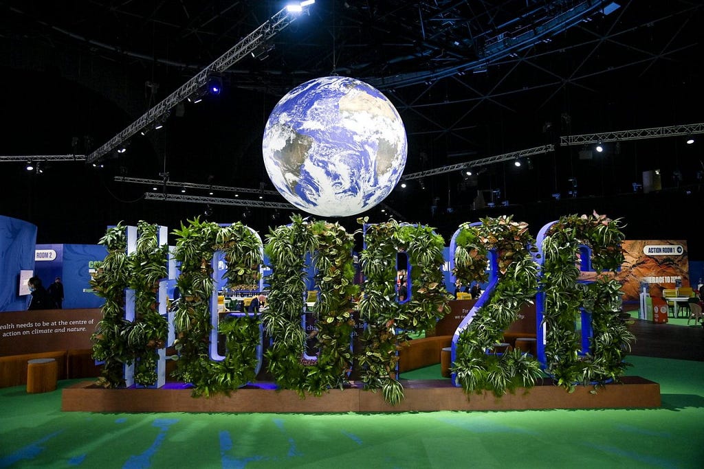 Model of a globe of Planet Earth suspended from the ceiling at the Glasgow COP26 UN Climate Conference, suspended above a large 3D rendition of the hashtag #COP26, which is covered in vegetation.