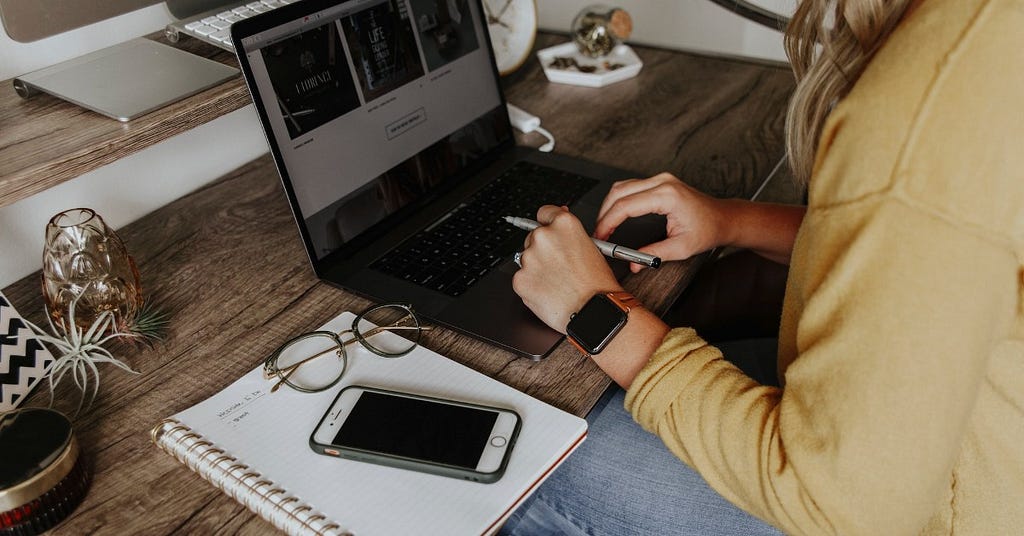 Woman at laptop computer with pen in her hand, notebook, and cell phone.