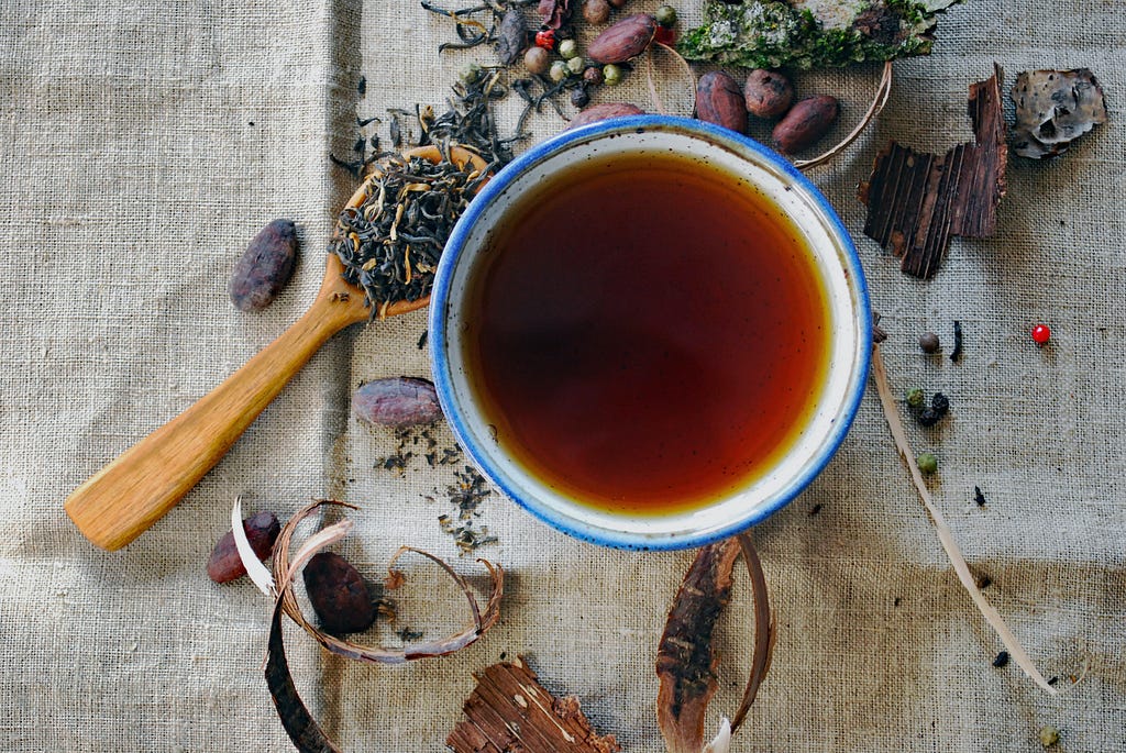 A top-down shot of a cup of black tea in a white mug. Strewn around it on a burlap table cloth are an assortment of loose tea leaves, including lavender. Some of these leaves are heaped into a small wooden spoon to the right of the cup of tea.