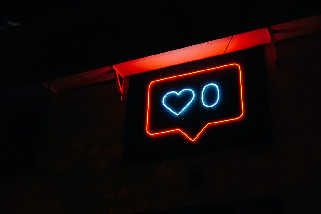 A red neon sign depicting a chatbox with a blue heart next to the number zero stands out against an otherwise dark background, with the exception of what looks to be a ledge that is being lit from the sign