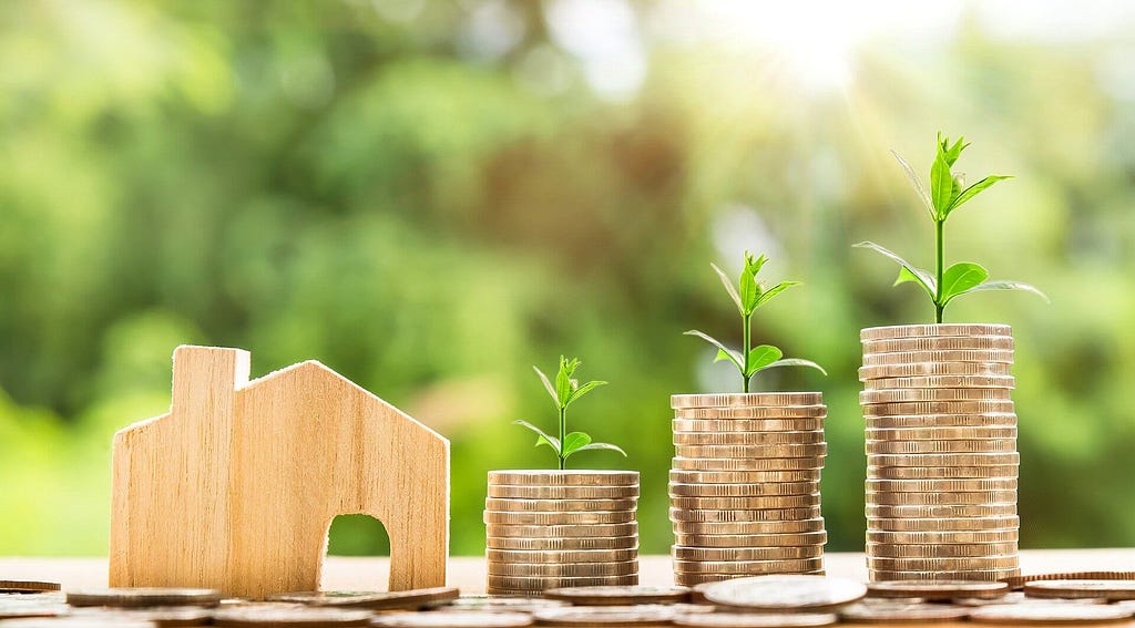 Wooden carved house sitting to the left of three progressively taller stacks of money coins with green leaves sprouting