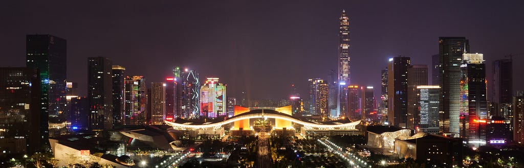 The cityscape of Shenzhen at night, with a purple black background sky and buildings with white and yellow lights.