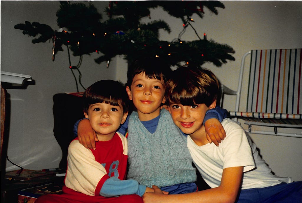 Three boys sit in front a sparely decorated Christmas Tree. The middle boy supports himself on the shoulders of the other two