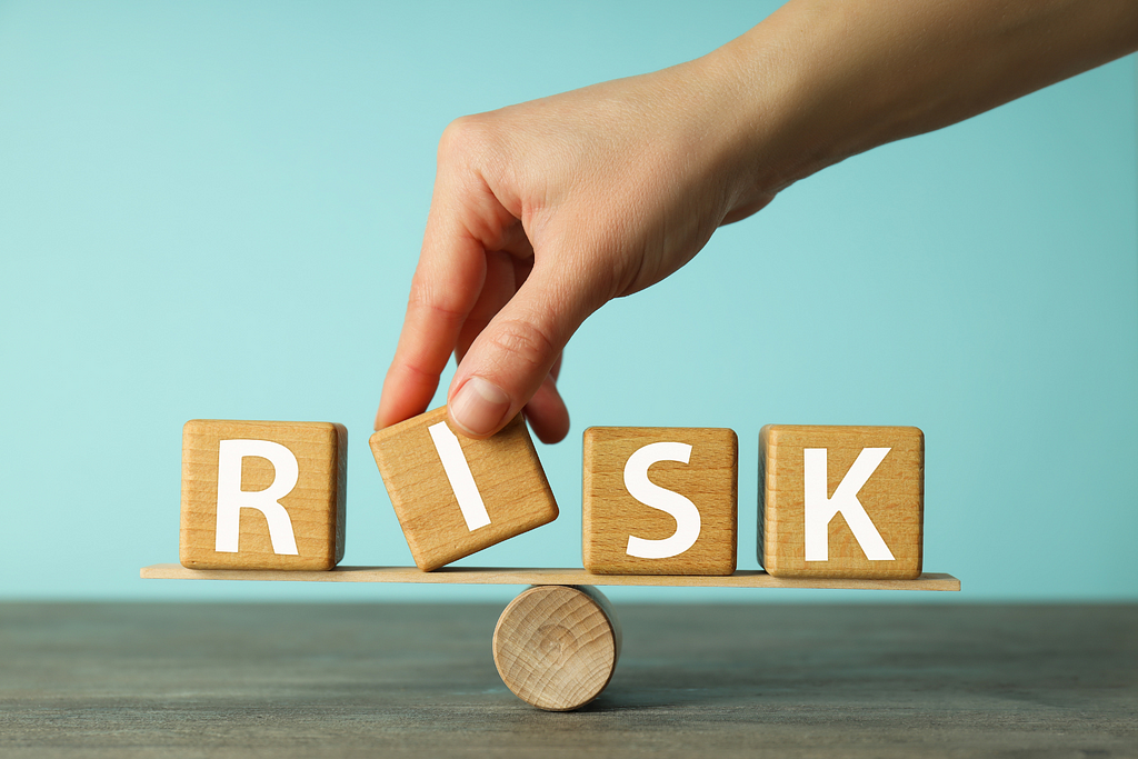 A hand carefully balances wooden blocks spelling out “RISK” on a seesaw, highlighting the concept of risk management.