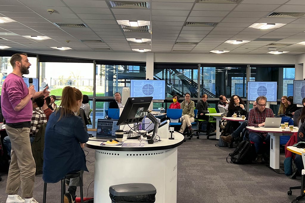 Professor Tim Heaton shares ideas for the Interdisciplinary Network on Time at their collaborative lab. He is standing to the left of the image in a brightly-lit room, with people sitting grouped around tables to listen.