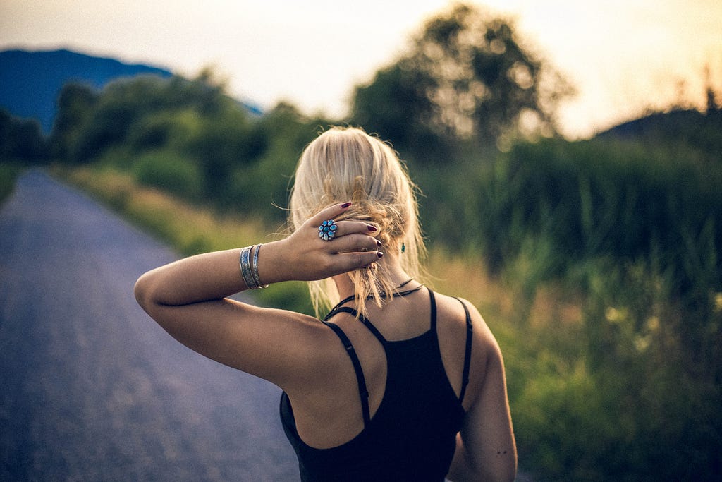 The sun sets behind a woman with blonde hair. Her back is to the camera. Her hair is pulled up in a loose bun. She’s wearing a black tank top.