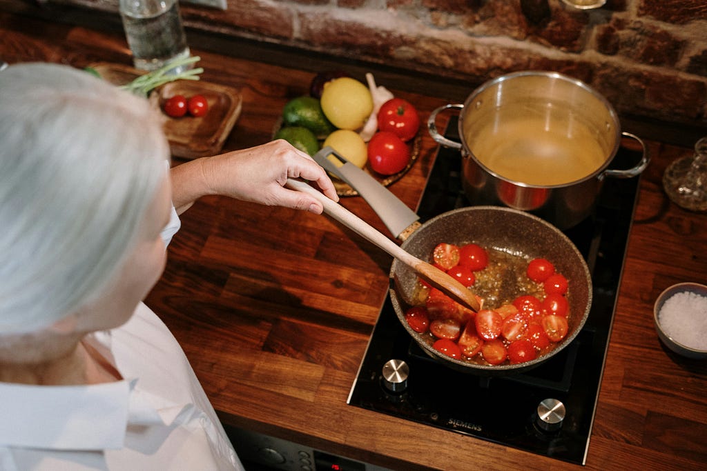 Older woman with grey hair cooking tomato and stock on hob.