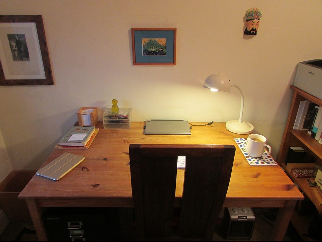 photograph of a wooden desk with chair pulled up to it. On the desk are a laptop, tablet, a mug, a desk lamp, 2 notebooks and a filing tray. There are two pictures on the wall behind the desk and alongside a mask.