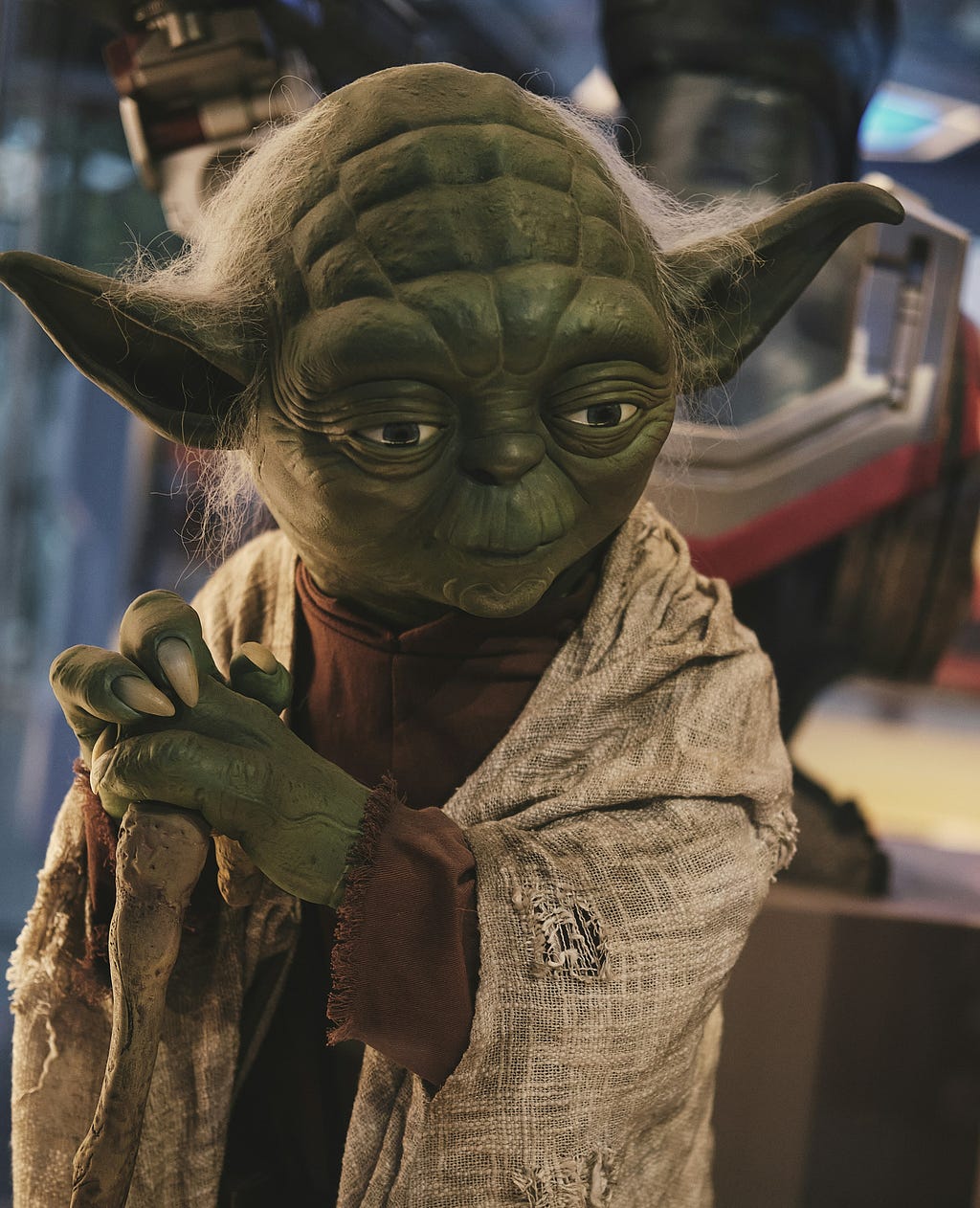Life-sized Yoda figure with detailed green skin, wrinkled features, and wispy gray hair, wearing his signature beige robe and holding a wooden walking stick.