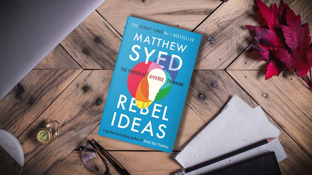 A copy of Matthew Syed’s Rebel Ideas lies on a parquet floor surrounded by intellectual paraphernalia like pencils, eye-glasses, laptops and paper