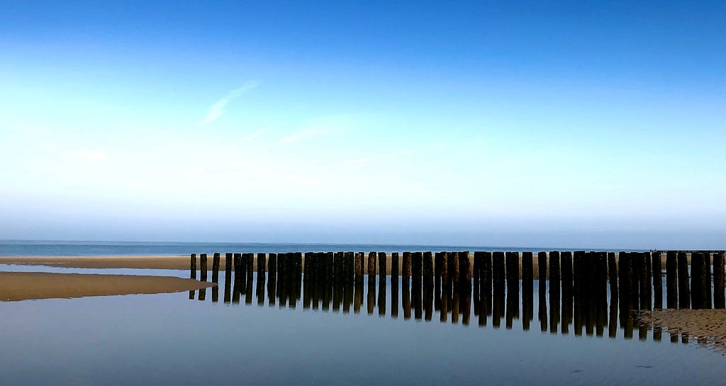 Landscape of Burgh-Haamstede, in the Netherlands, depicting a sandy beach with sea in the distance and shallow water in the foreground. There is a line of posts — wave breakers — driven into the sand.