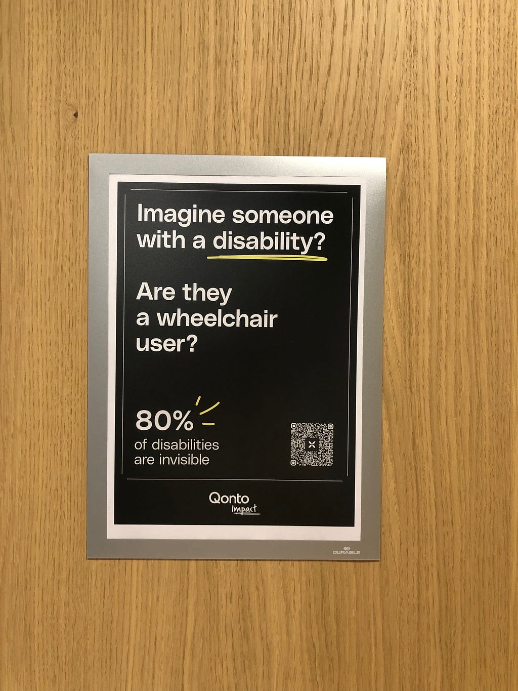 Poster on an office wall with an eye-catching headline: “Imagine someone with a disability”. The next line reads: “Are they a wheelchair user?” The final line reads: “80% of disabilities are invisible”. The poster is signed “Qonto Impact”.