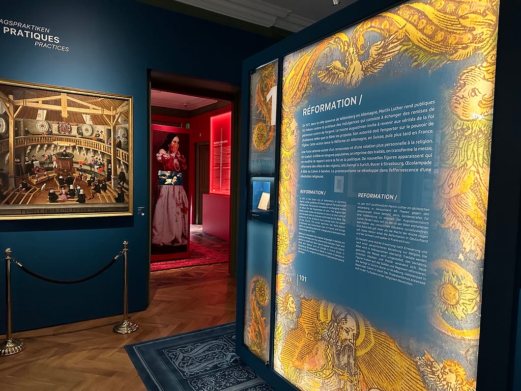 An exhibition space with deep blue walls, showcasing religious history. The left displays a painting and the right features a large informational panel with golden textile patterns framing the text on Reformation