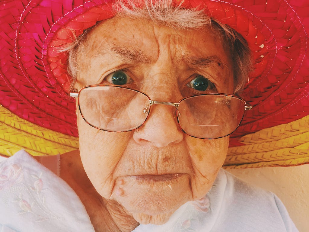 The face of an elderly woman, wearing glasses on her nose and a wide brimmed straw hat