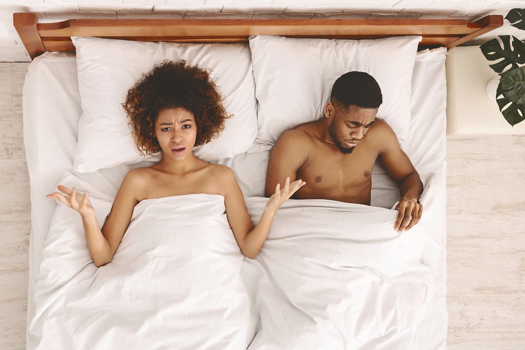 Sex problem. Disappointed black unsatisfied woman lying in bed, sad black man looking under blanket, top view.