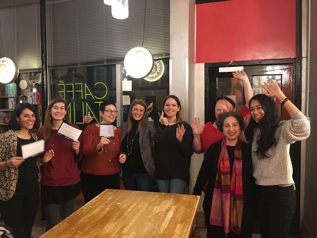 Photo of 8 women smiling, some holding up cards, some waving at camera. In a cafe, standing up behind table.