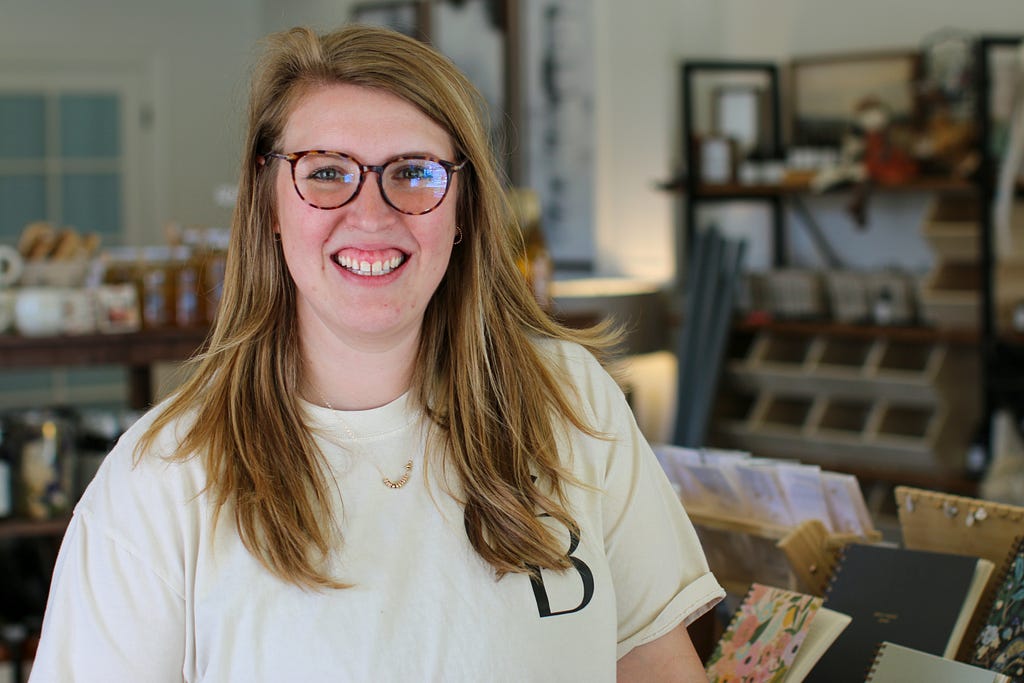 A woman with long hair, glasses and wearing a t-shirt stands in a retail space, smiling at the camera. Shelves and products are visible in the background