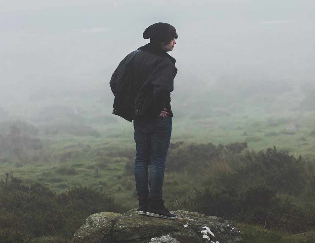 Photo of a man in a black hat and black coat and jeans standing on a ledge looking out over a foggy field