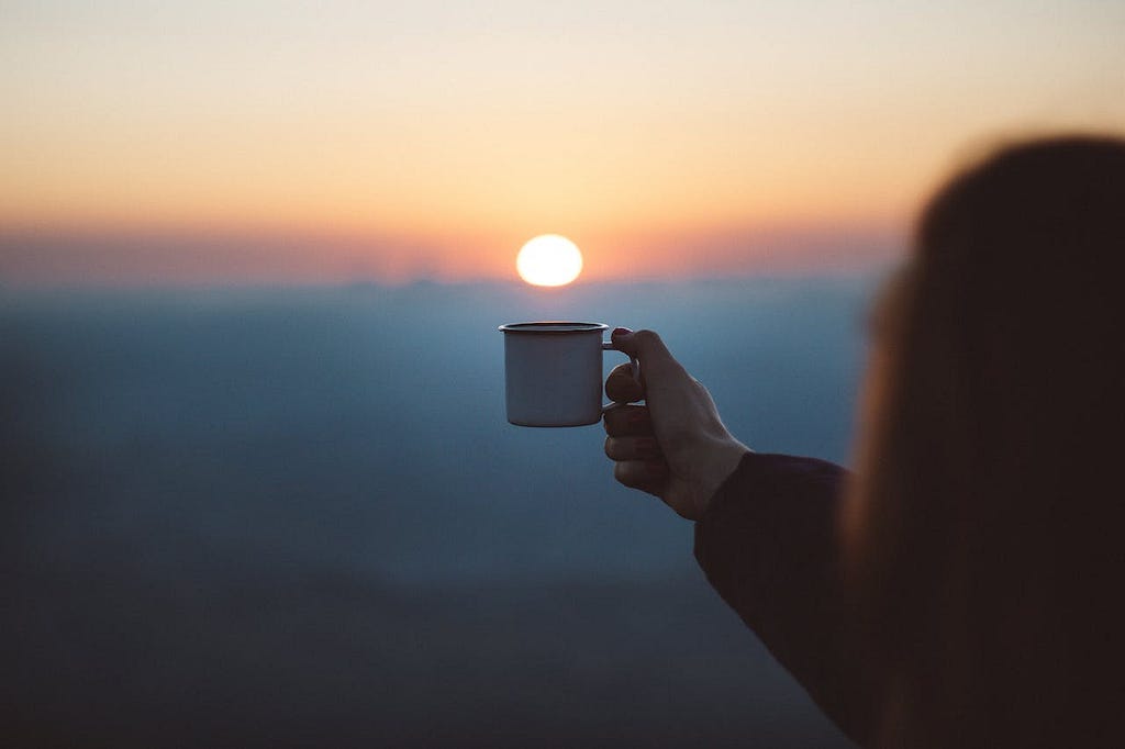 A woman holding up a coffee mug to align with the setting sun in the sky so it appears that the sun is setting into the mug.