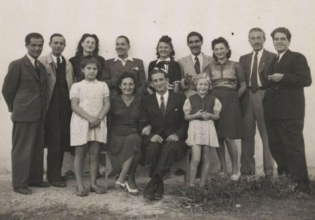 A black-and-white group portrait. In the front row, two young girls in white dresses stand on either side of Karl and Ella Schwarz. Karl is wearing a dark suit and a tie, and Ella is wearing a dark two-piece skirt suit and white shoes. Nine smiling adult men and women stand behind them, also in dresses and full suits.