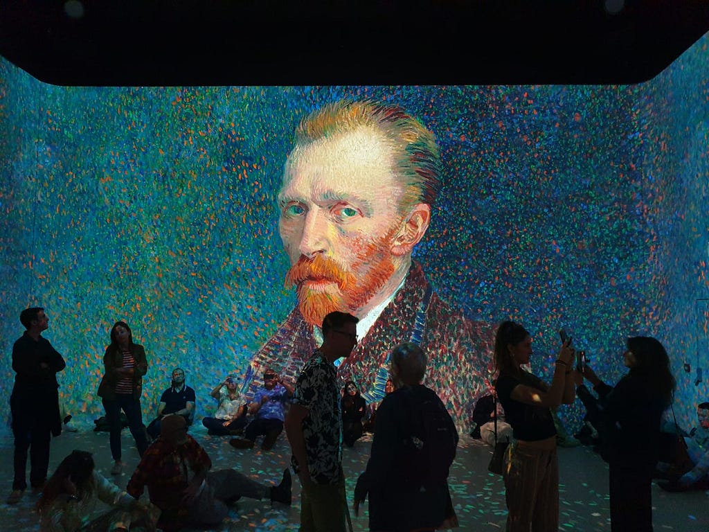 Visitors stand silhouetted against a large-scale rendering of Van Gogh’s Self-portrait. A blackened void above the screen is visible