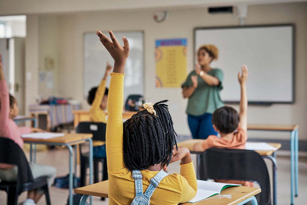 Classroom with a Black woman teacher at the front, and children raising their hands in the desks. Photo by pixdeluxe/Getty Images