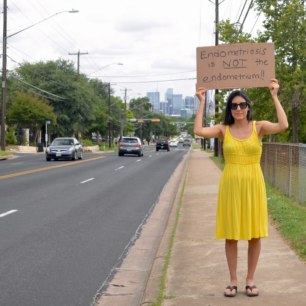 Amy is in a yellow dress standing on the sidewalk of a busy road with cars passing by. She holds a sign over her head that says ‘Endometriosis is not the endometrium.’