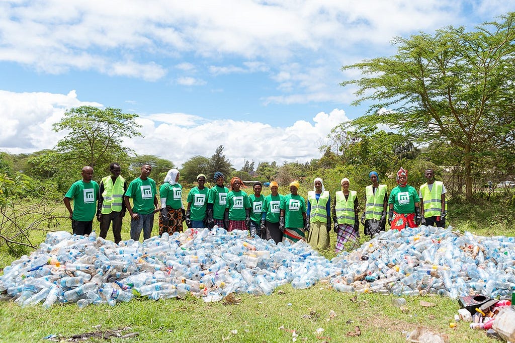 A photo of fifteen participants of a cleanup activity in Tanzania, and the large mounds of plastic bottles and other waste they have collected for recycling.