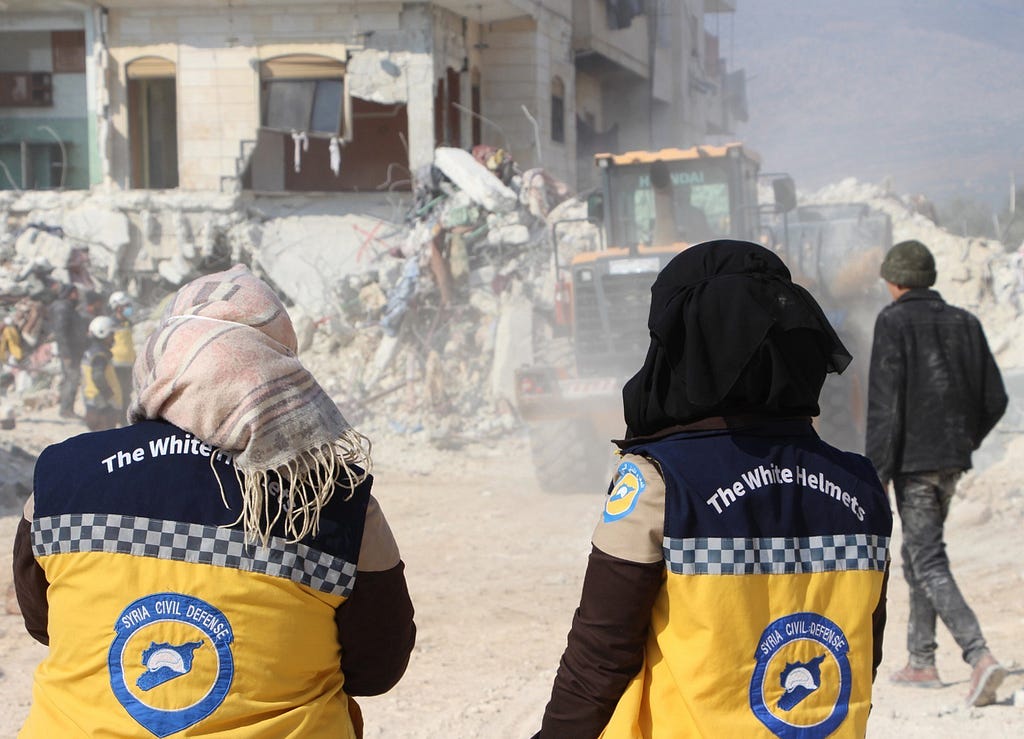 Two women face away from the camera. They wear yellow and black White Helmets vests and head coverings. In front of them are piles of rubble and damaged building. A yellow and black construction vehicle is at the center of the scene. In the left background, a group of people wear white helmets. At right, a person wearing black walks into the frame.