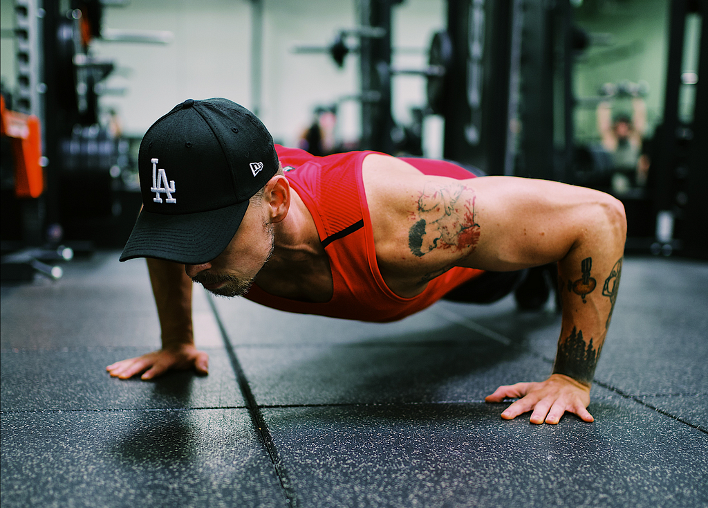 Man in gym wearing a red tank top and black cap, doing a pushup