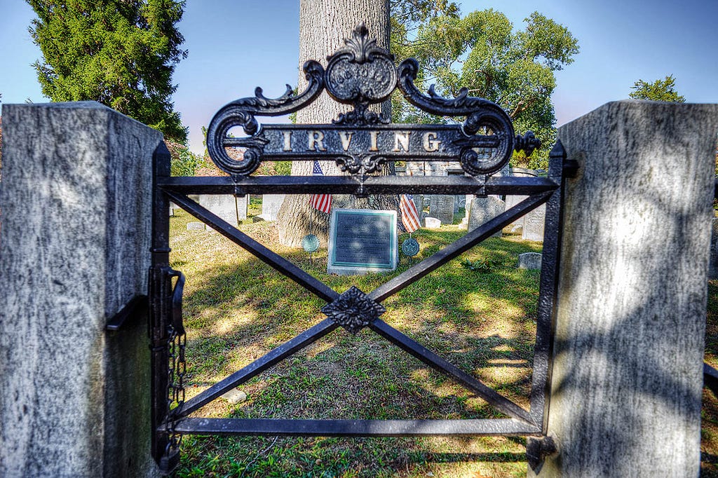 An ornate wrought iron gate with swirling designs partially conceals a pathway leading into a cemetery.