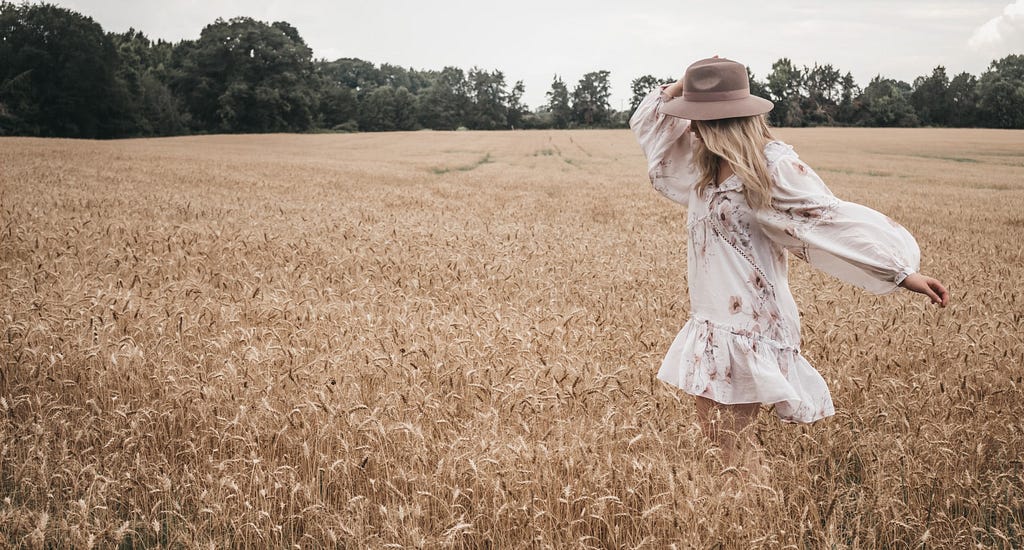 A woman with long blonde hair is walking alone in a large field. She’s wearing a hat and flowy dress. The hat and her hair obscure her face and she appears to be focused inward.