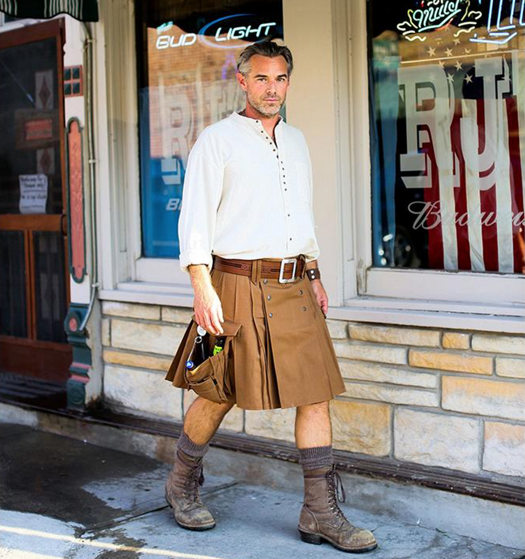 White, masculine person walking down sidewalk near a beer store staring at camera, with white buttoned shirt with brown kilt with large brown belt and open side pocket containing beer bottles. Wearing socks and worn brown high boots.
