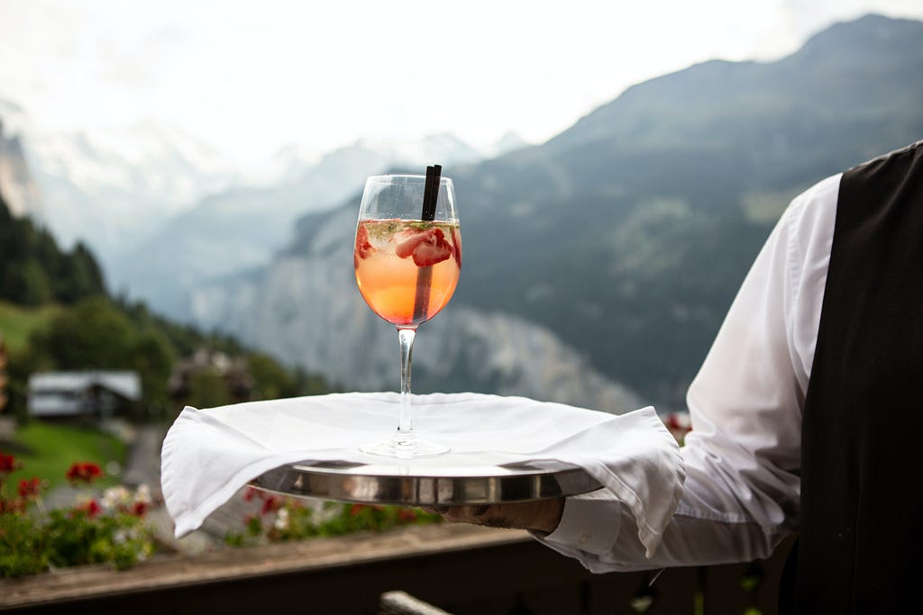 A food service worker offers a drink in a long-stemmed glass, overlooking a picturesque view of mountains.