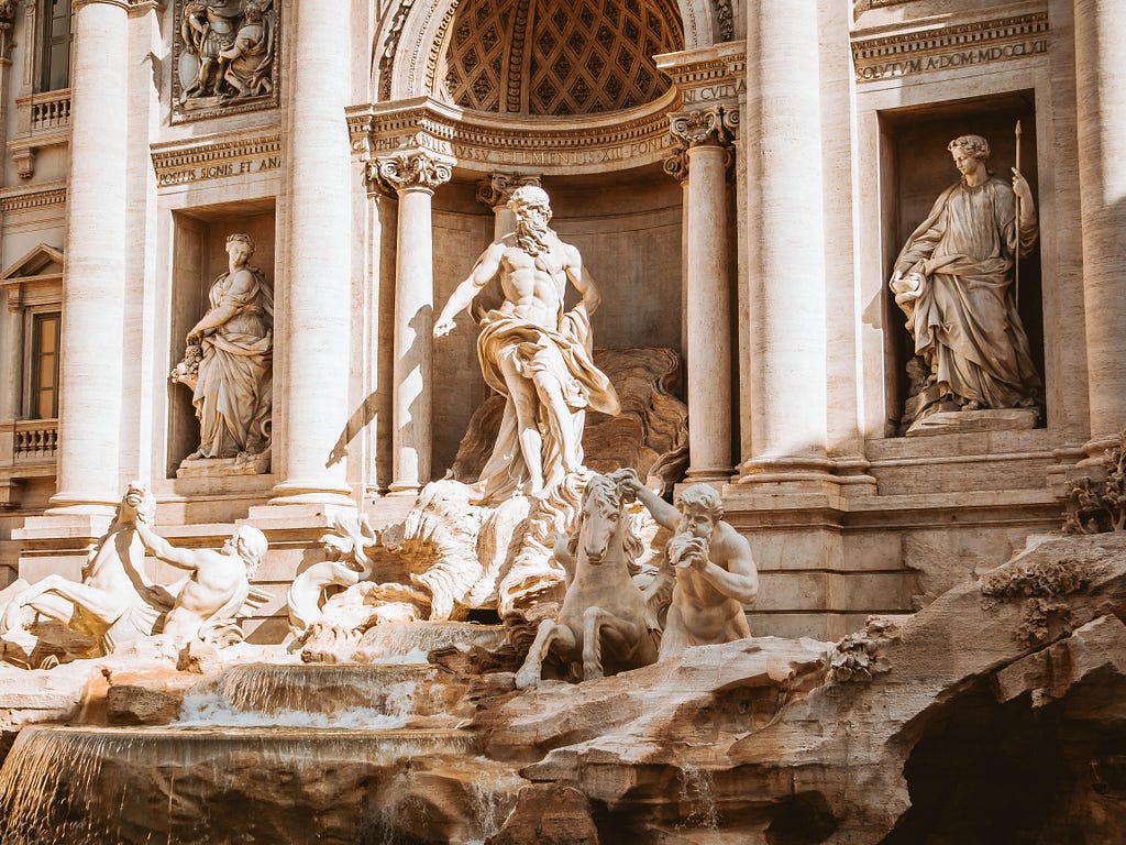 A view of the Trevi Fountain. Sculptures of roman gods and horses sculpted in white marble are touched by gentle ray of sun.