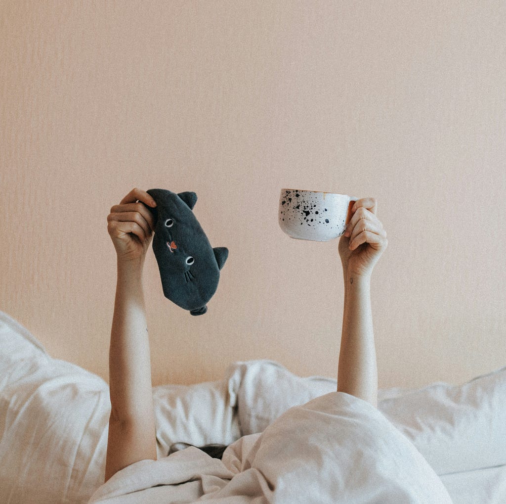 A person lying in a bed, covered head to toe with a white duvet. They are raising their arms, holding a black cat sleep mask in one hand, and a white ceramic mug in the other.