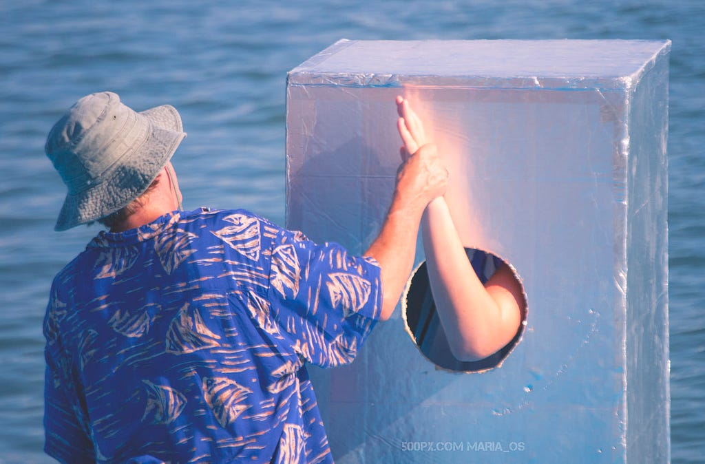 A man in summer shirt and hat holding other man’s hand, concealed in the box