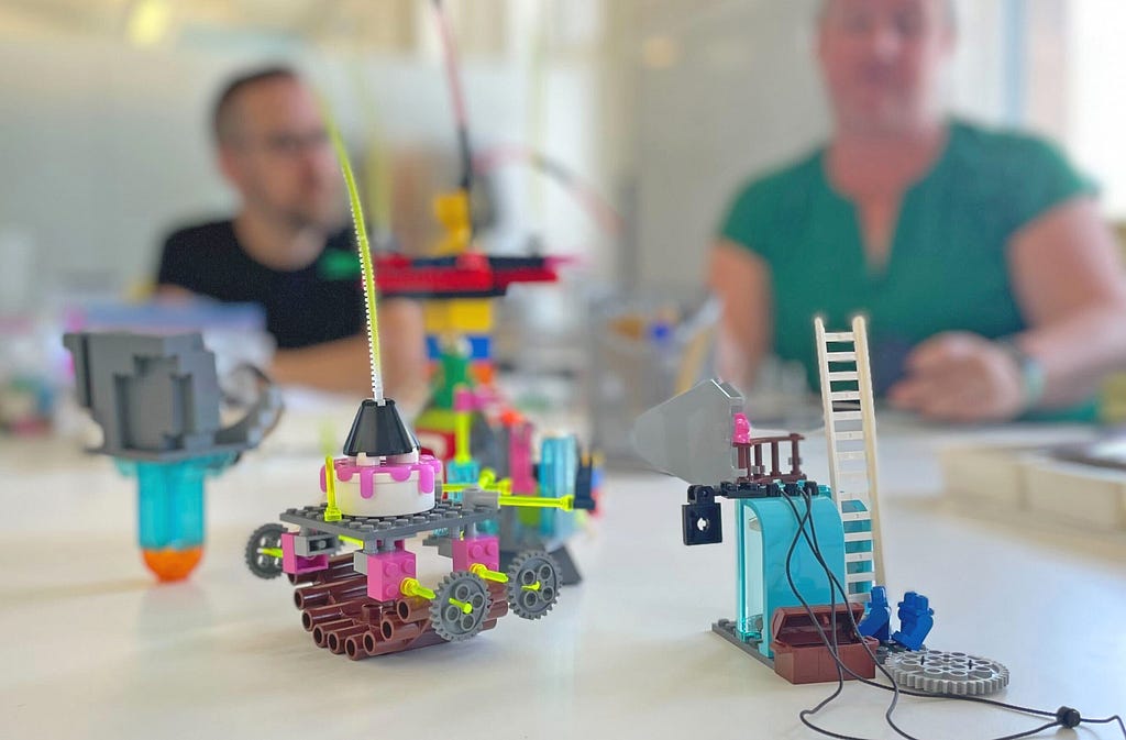 A closeup of several participant’s ambiguous but detailed LEGO models in the foreground, while blurred in the background is Nick taking part while Andrea facilitates