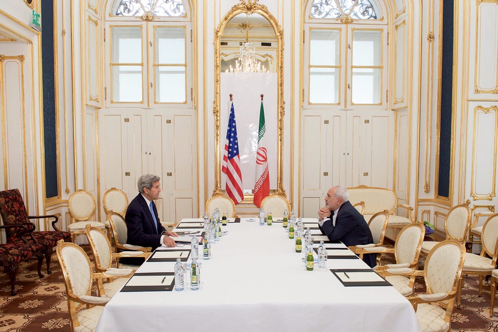 John Kerry and Javad Zarif sit across a table in front of American and Iranian flags in the Blue Salon of the Palais Coburg in Vienna.