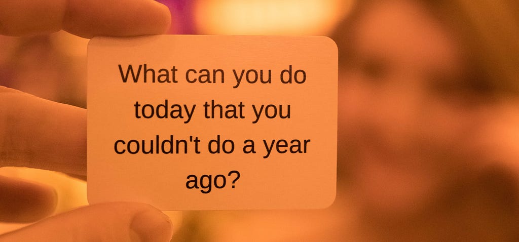A closeup of someone holding a small sign saying ‘What can you do today that you couldn’t do a year ago?’.