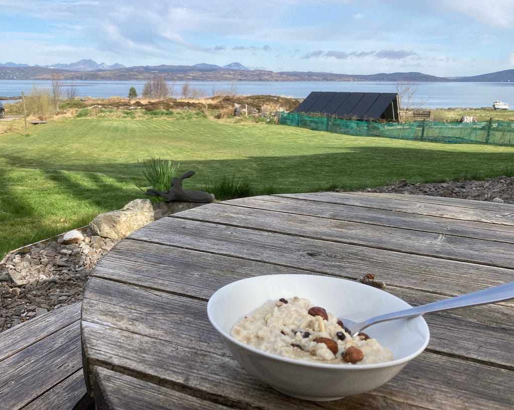 A bowl of porridge sits outside on a wooden table. In the distance is beautiful Scottish landscape of hills and water