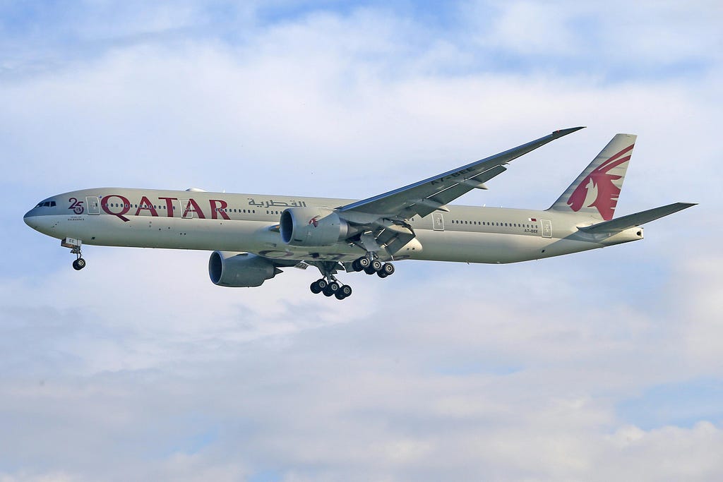 The image shows a Qatar Airways Boeing 777 aircraft in flight against a backdrop of a partly cloudy sky. The aircraft is painted in the airline’s distinctive livery, featuring the word “QATAR” in large maroon letters on the fuselage and the Oryx logo on the tail. The plane’s engines and landing gear are visible, and it appears to be in the process of landing or taking off. The number “25” is also visible near the front, likely indicating a special marking for Qatar Airways’ 25th anniversary. The