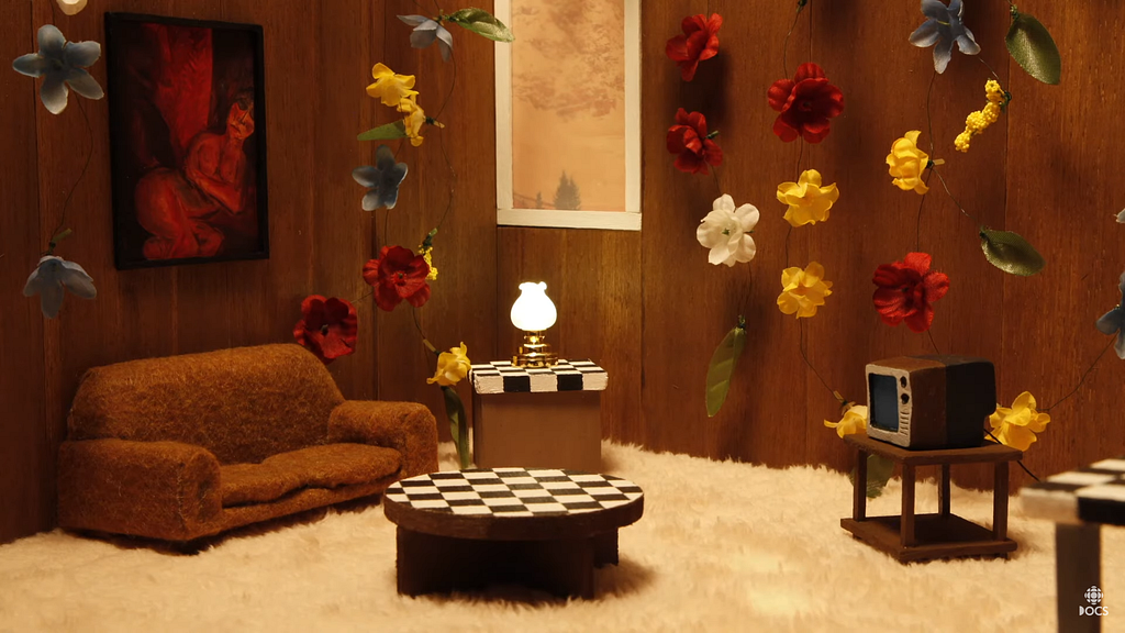 Still from IN THE SHADOW OF THE PINES. Diorama setting of a white furry carpeted living room with a brown couch on the left, checkerboard coffee table and side table with a lit lamp sitting on it, and a television set on the right. There are paintings hung on the walls behind the couch and the side table, and flowers are hanging down on all the other walls.