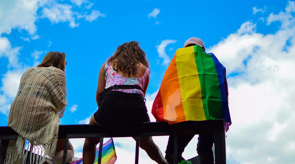 Three figures sit on bleachers, their backs to the camera. Behind them is all blue sky and clouds. The figure on the right has a rainbow flag wrapped around him