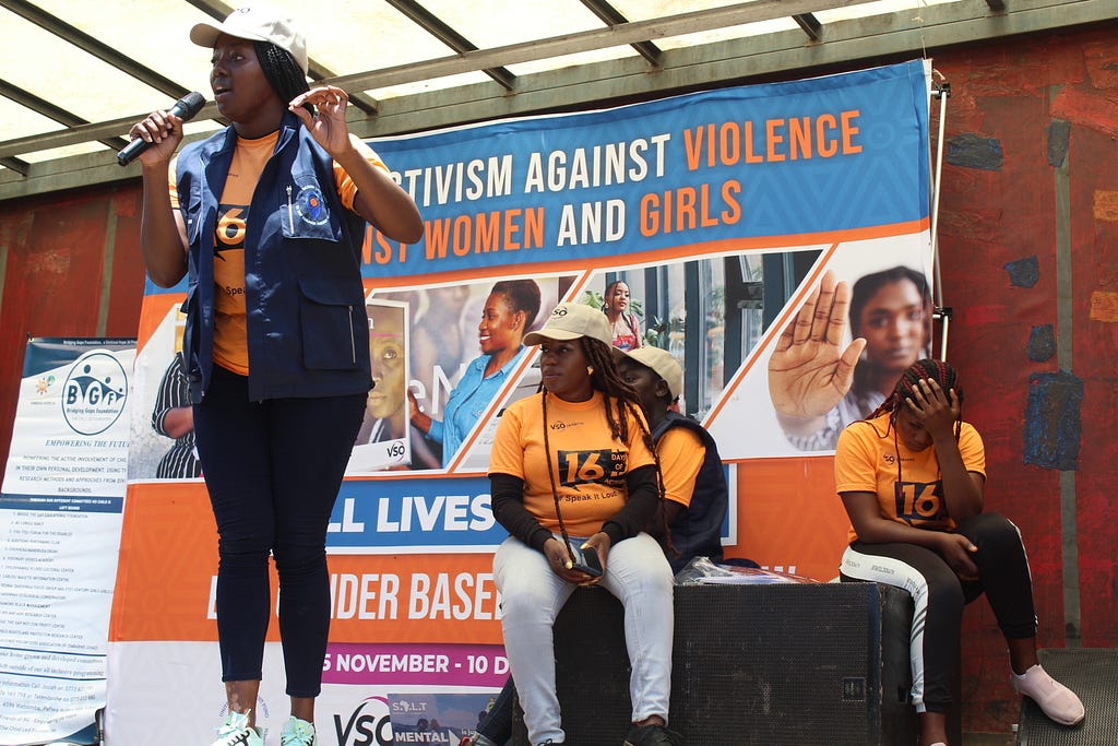 A woman wearing an orange t-shirt is standing on stage, speaking into a microhpne. In the background, we can see a banner that reads “Activism against violence against women and girls”, and one woman and one man are sitting.