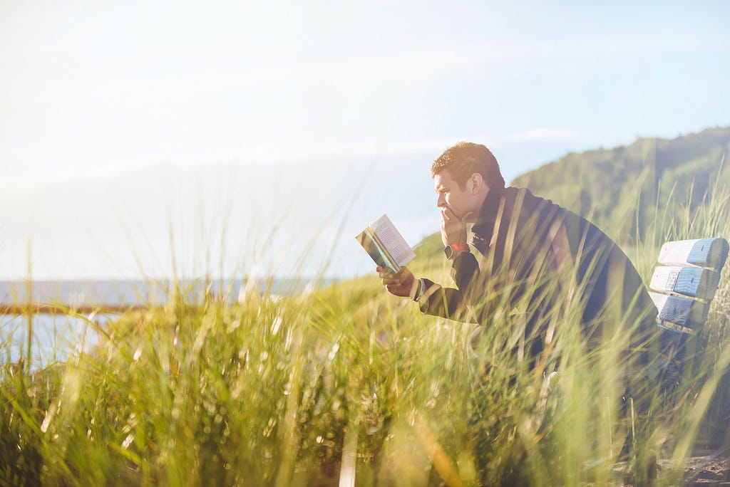 man reading book and gaining knowledge in a field overlooking a lake with mountains in the distance