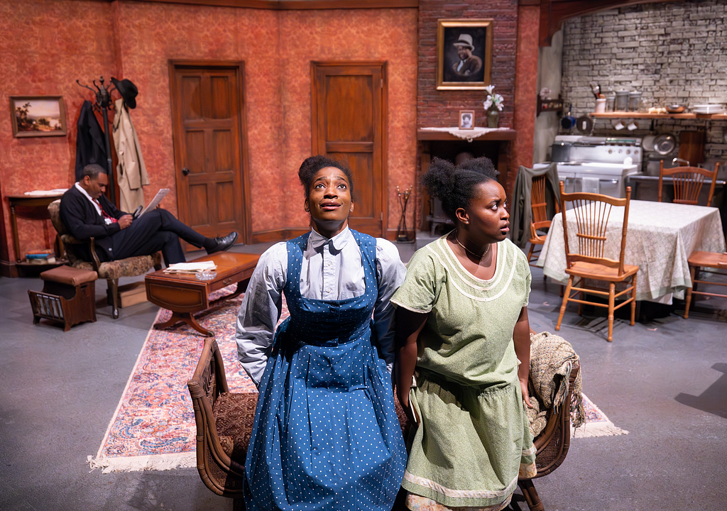 Two girls in 1950s dresses sit at the front of the stage while a man sits and reads the paper behind them. The actress on the left looks up excitedly while the one on the right looks to the side apprehensively.