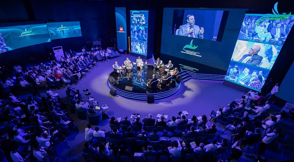 Panelists sit in the middle of a crowded room with digital screens in the background.