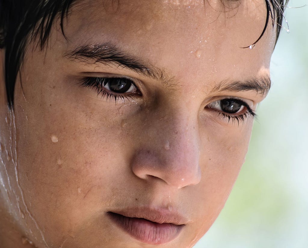 The face of a nine-year-old Venezuelan boy tipped downwards. The sun shines on his face and highlights wisps of wet hair and droplets of water trickling down his face